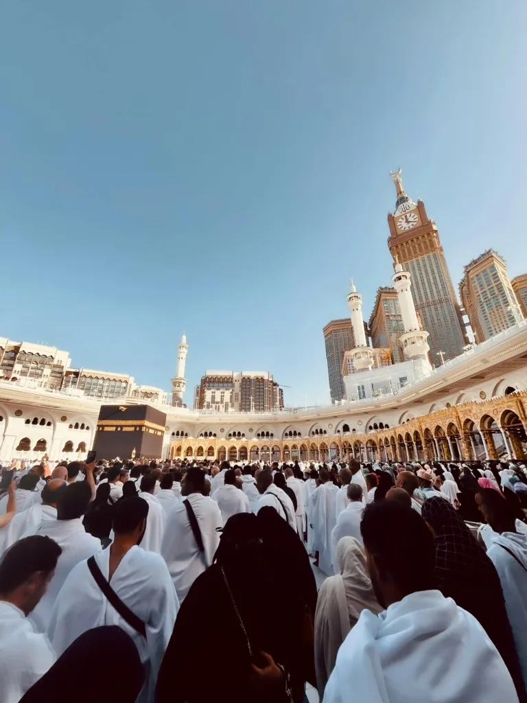 Kerumunan jemaah haji berpakaian ihram memadati Masjidil Haram dengan latar belakang Menara Jam, merepresentasikan niat suci dalam menentukan prioritas apakah ingin menghajikan orang tua atau diri sendiri terlebih dahulu bersama Duha Wisata.
